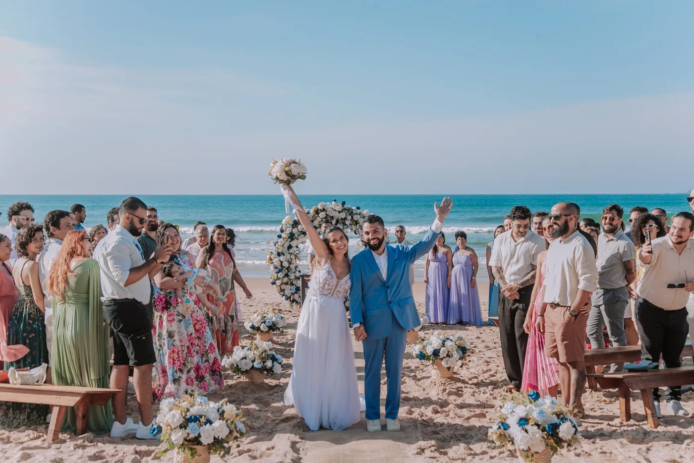 Casal comemorando com os braços para o alto ao fim da cerimônia na praia, rodeado pelos convidados