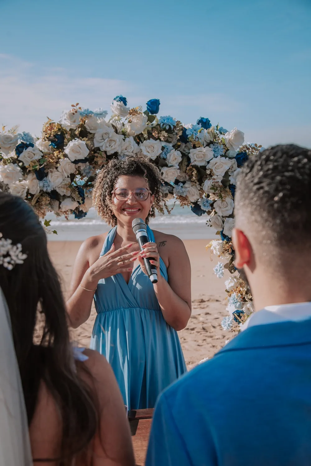 Fabiana celebrando uma cerimônia de casamento diante de um arco de flores, com os noivos de costas em primeiro plano
